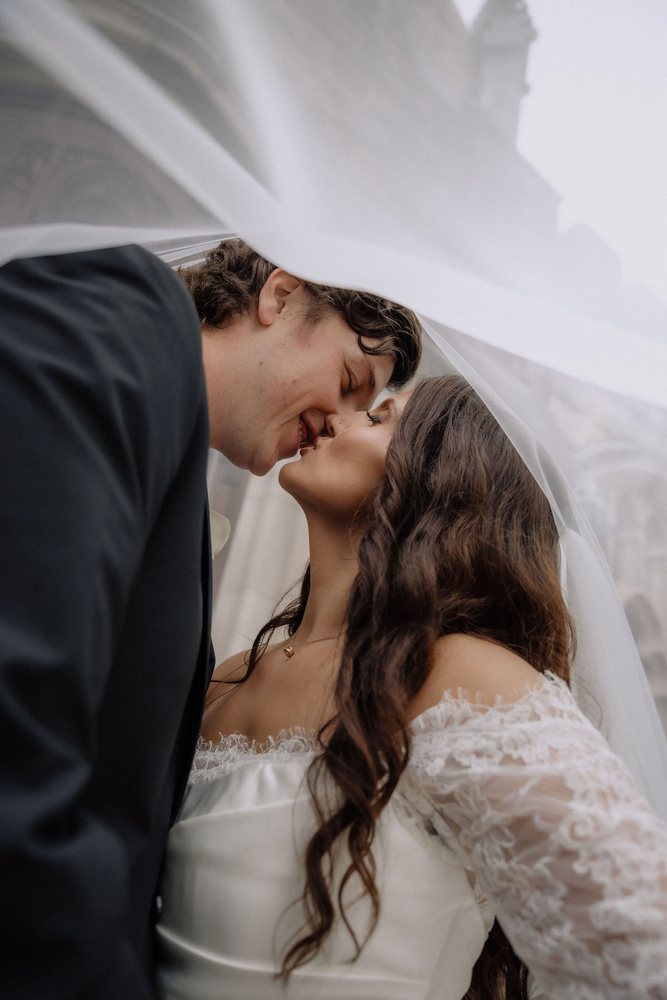 A bride and groom kissing underneath her veil outside a stone chapel