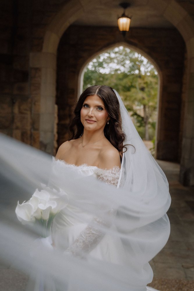 A bride standing under a stone archway with her veil swirling around her