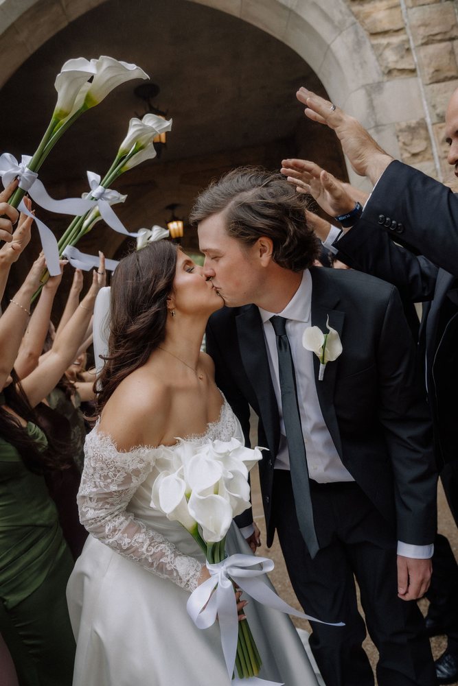 A bride and groom kissing as guests raise calla lilies overhead on their way out of the chapel