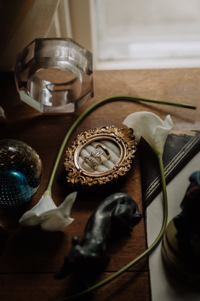Wedding rings resting in an ornate gold frame beside calla lilies on a dark wood table