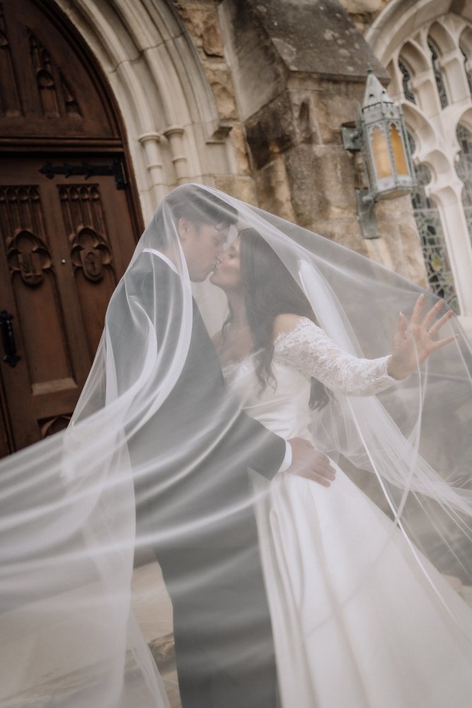 A couple sheltered under a sweeping cathedral veil at the doors of a stone chapel
