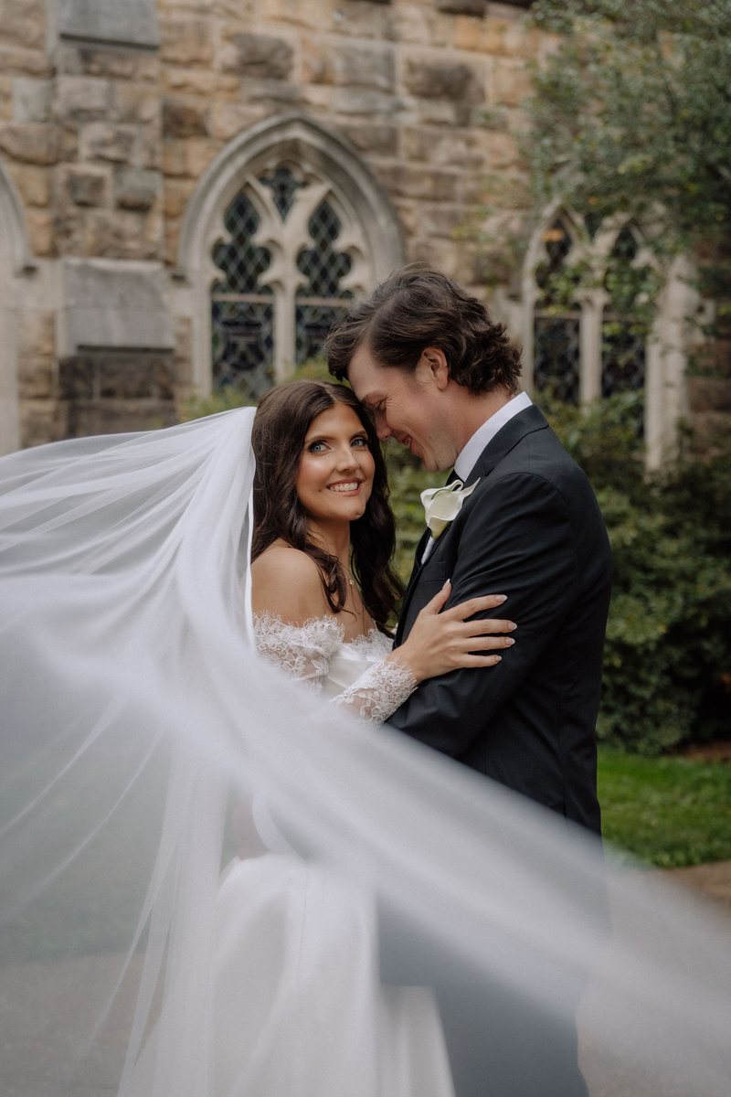 Lexi and Mason with her veil sweeping across the frame outside All Saints Chapel