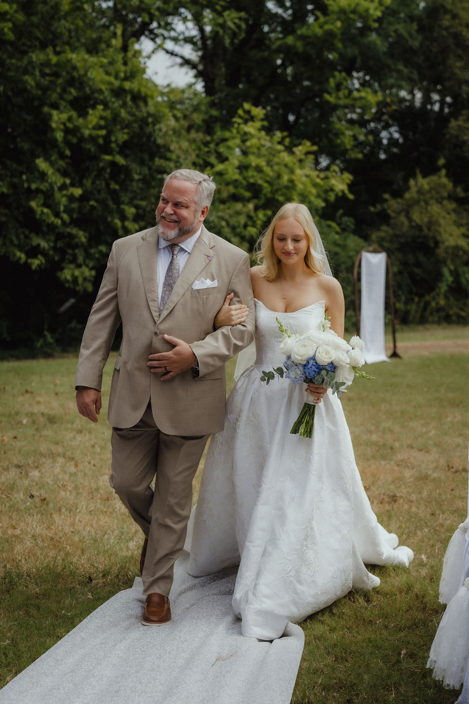 Denalyn coming down the grass aisle on her father's arm, her train out behind her