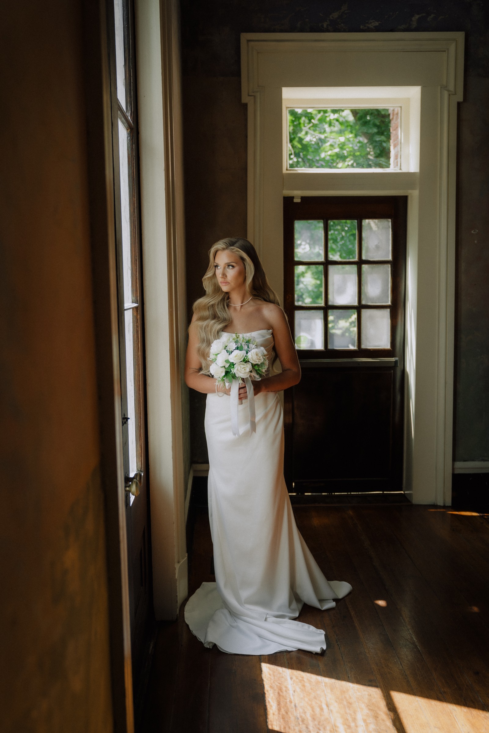 Maddie in the hallway doorway with her bouquet, light spilling across the floor