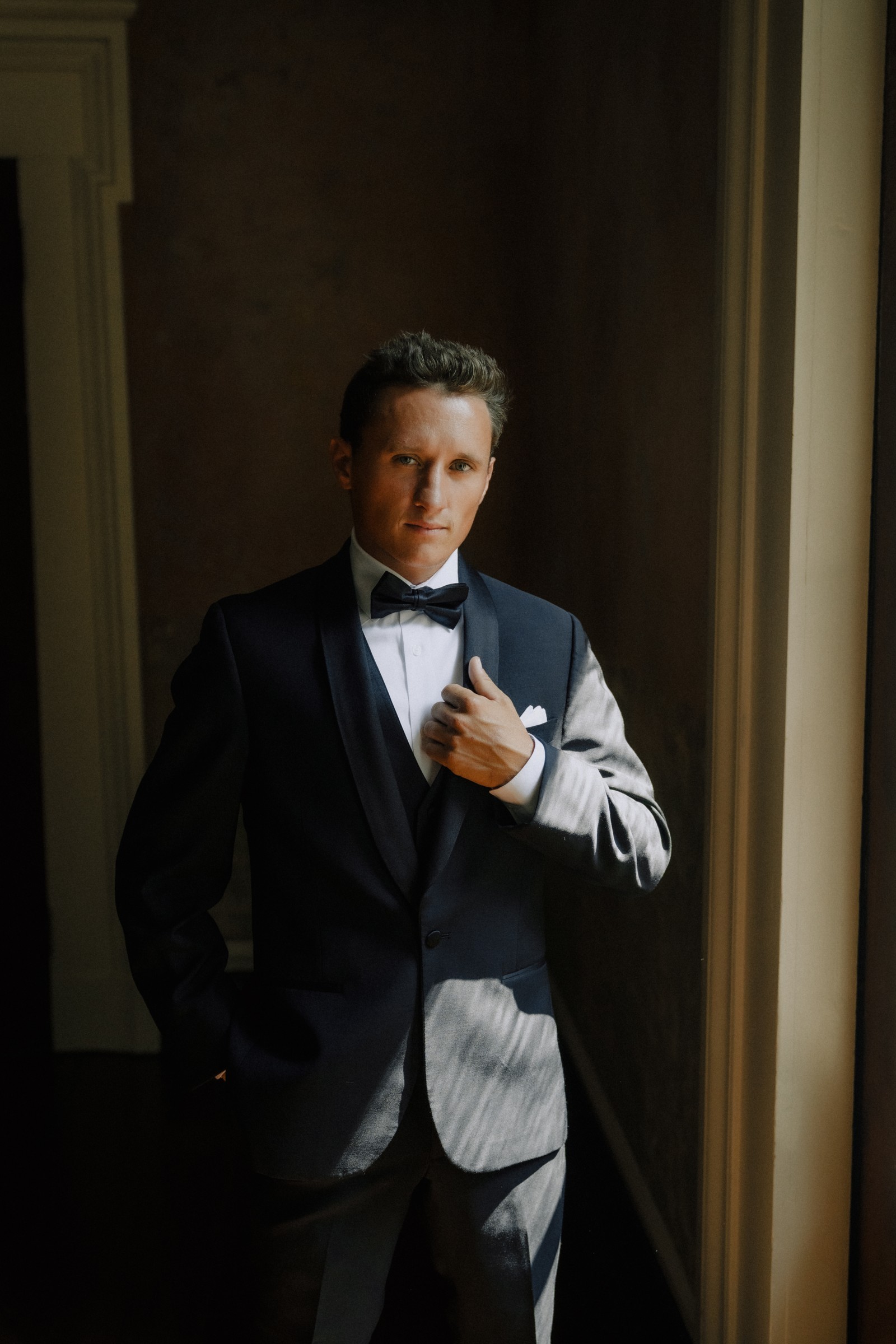 Brandon adjusting his tux in a hard slash of window light