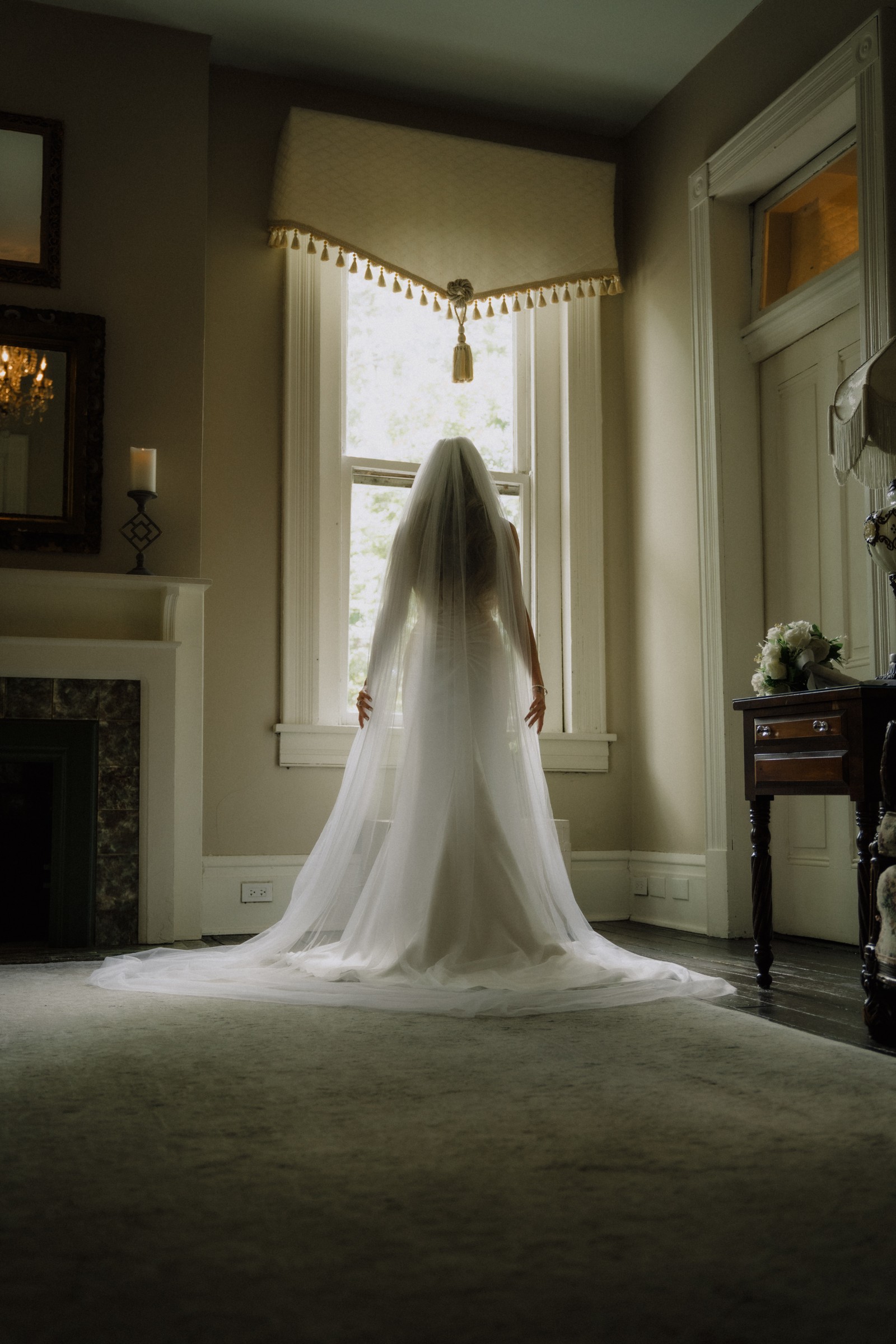 Maddie facing the window, her veil held out like wings, bouquet waiting on the side table